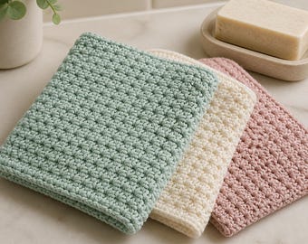 May include: Three square crocheted washcloths stacked together in sage green, cream, and dusty rose colours. Each cloth features a consistent, textured star-like stitch pattern. The items are arranged neatly, showcasing the yarn texture and colour variations.