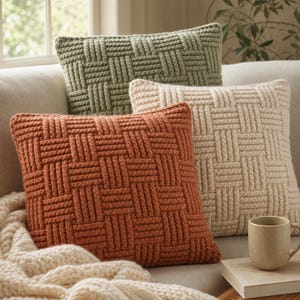 Può includere: Tre cuscini quadrati caratterizzati da un motivo a uncinetto con trama a intreccio in verde salvia, crema e arancione bruciato. I cuscini sono disposti insieme, con una spessa coperta in maglia grossa color crema drappeggiata in primo piano.