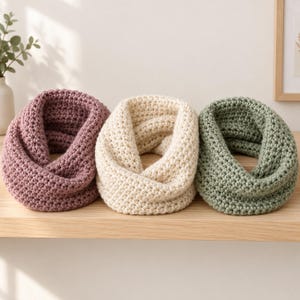 May include: Three chunky infinity scarves arranged in a row. The left scarf is a dusty mauve, the center scarf is a creamy off-white, and the right scarf is a muted sage green. Each scarf features a thick, textured crochet knit pattern.