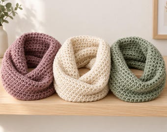 May include: Three chunky infinity scarves arranged in a row. The left scarf is a dusty mauve, the centre scarf is a creamy off-white, and the right scarf is a muted sage green. Each scarf features a thick, textured crochet knit pattern.