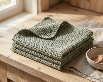 May include: A stack of three sage green square cloths featuring a textured, waffle-like crochet stitch pattern. The cloths are folded neatly, displaying a consistent, uniform weave throughout the fabric, with a soft, matte finish on the surface.