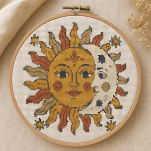 May include: A circular wooden embroidery hoop containing a cross-stitch design of a sun and moon face. The sun is yellow with orange and brown rays, while the moon is white with dark blue accents. Small stars surround the central celestial motif.