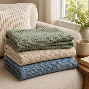 Puede incluir: Una pila de tres mantas dobladas con un patrón de tejido de punto tipo gofre. La manta superior es de color verde salvia, la del medio es beige claro y la inferior es de un tono azul apagado. Cada manta está doblada en forma rectangular.