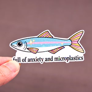 May include: A sticker featuring a cartoon fish with a light blue body, pink and yellow fins, and the text "full of anxiety and microplastics." The fish has a large eye and star accents.
