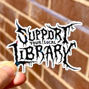 Puede incluir: Una pegatina con el texto "SUPPORT YOUR LOCAL LIBRARY" en una fuente negra de estilo gótico. La pegatina tiene un borde blanco y acentos iridiscentes. El fondo es una pared de ladrillo.