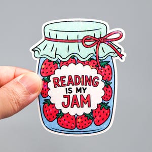 Può includere: Un adesivo raffigurante un barattolo pieno di fragole. Il barattolo ha un coperchio azzurro e un fiocco rosso. Il testo "READING IS MY JAM" è in una bolla bianca al centro del barattolo.