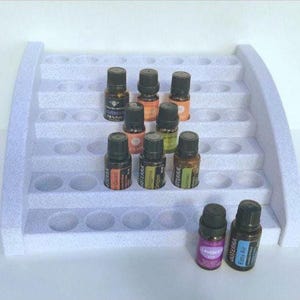 May include: A white, tiered foam display stand with multiple circular cutouts for organising small bottles. Several dark glass essential oil bottles with labels are arranged on the steps and at the base of the stand, featuring various coloured caps.
