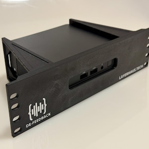 Puede incluir: Dispositivo electrónico negro con el texto "LAYERHAUS TECH" y "DE-FEEDBACK". El dispositivo tiene forma rectangular con múltiples puertos y orificios de ventilación. El panel frontal tiene un logotipo con un soporte y un diseño de onda de sonido.