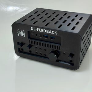 May include: Black electronic device with the text "DE-FEEDBACK" and a graphic. The device has multiple buttons, knobs, and ports. The top of the device has a series of rectangular cutouts for ventilation. The device is sitting on a white surface.