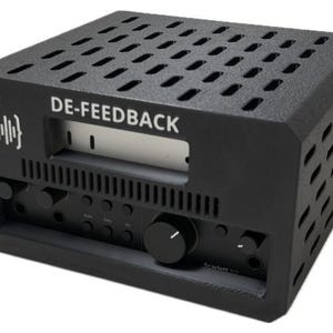 May include: A black audio interface enclosure labelled DE-FEEDBACK with a white sound wave icon. The rectangular housing features a perforated top and side panels for ventilation, with a recessed front panel containing control knobs and buttons.