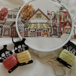 May include: Cross-stitch embroidery in a white hoop, depicting a detailed village scene with autumn colors. Three thread bobbins with black cat-shaped holders and thread colors of red, yellow, and green. Gold scissors are also present.
