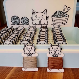 Op de afbeelding: Een lichtblauwe opbergdoos met talloze witte plastic borduurgarenklosjes in de vorm van kattenkoppen. Het deksel is voorzien van zwarte lijntekeningen van garenbollen, een kat en een mand met breinaalden. Twee klosjes met 646 en 433 staan ervoor.