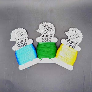 May include: Three spools of embroidery floss in blue, green, and yellow, each wrapped around a white hedgehog-shaped holder. The holders have the numbers 996, 699, and 726 printed on them. The background is a solid gray.
