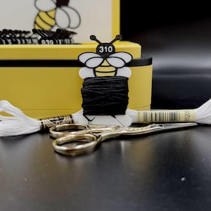 May include: A black embroidery thread wound on a bee-shaped plastic bobbin labeled 310, positioned in front of a yellow storage box. A skein of white embroidery thread and a pair of gold-toned metal scissors are placed in the foreground.
