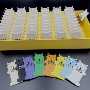 May include: A collection of cat-shaped tags in a yellow tray, with several colorful cat tags in front. The tags are white, black, purple, green, orange, yellow, light blue, and white. Each tag has the number "223" printed on it.