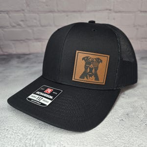May include: Black trucker hat with a mesh back and a curved brim. The hat features a brown leather patch with a dog wearing sunglasses. The patch is square and centered on the front of the hat. The hat is adjustable and has the Richardson brand label.