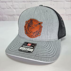 May include: A heather gray trucker hat with a black mesh back and a circular orange leather patch featuring a marlin graphic secured by four metal rivets. The brim displays a black sticker with the text Richardson Original 112 Trucker Adjustable.