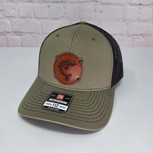 May include: An olive green trucker hat with a black mesh back and a brown circular leather patch featuring a black fish skeleton graphic. The front of the brim displays a black and white sticker with the text Richardson Original 112 Trucker.