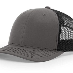 May include: A charcoal gray trucker hat with a black mesh back. The hat features a curved brim and a button on top. The front panel is solid gray, while the back is a breathable black mesh.