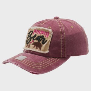 May include: A distressed maroon baseball cap featuring a rectangular patch on the front. The patch displays the embroidered text MAMA in pink and Bear in black, accompanied by a brown bear graphic and two small pink hearts above the bear.