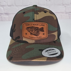 May include: A camouflage print snapback cap with a black mesh back. A rectangular tan leather patch is centered on the front, featuring an engraved fish graphic, two stars, and the text "TABLE ROCK LAKE MISSOURI". A circular sticker is on the brim.