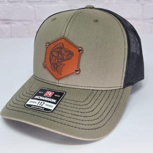 May include: An olive green trucker hat with a black mesh back and a brown hexagonal leather patch featuring a fish illustration. The patch is secured with four silver rivets. A black sticker on the brim reads Richardson 112 Trucker Adjustable.