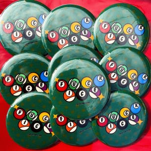 May include: A collection of green, round buttons with a pool ball design. Each button features the word "FUCK" spelled out with pool balls, and the letter "C" and "E" are also included. The buttons are arranged on a red background.