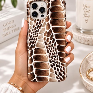 Puede incluir: Una funda para smartphone con un patrón de piel de serpiente en marrón y blanco con escamas de distintos tamaños. El diseño cubre toda la parte posterior de la funda, incluida la zona alrededor del recorte para la cámara de tres lentes.