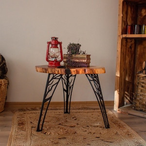 May include: A rustic, live-edge wooden side table with black metal legs. A red lantern sits atop the table, alongside a small stack of books and dried plants. A cat sits on a wicker basket to the left.