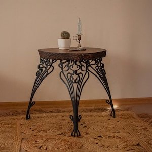 May include: A small, round side table with a dark, textured wooden top and ornate black metal legs. A small cactus in a white pot, a candle holder with a lit candle, and a small object rest on the table. The table is on a woven rug.