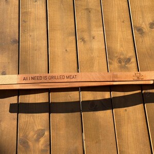 May include: Wooden barbecue tongs with the text "ALL I NEED IS GRILLED MEAT" engraved on the top. The tongs are made of light brown wood and feature a fox logo. The tongs are resting on a wooden surface.