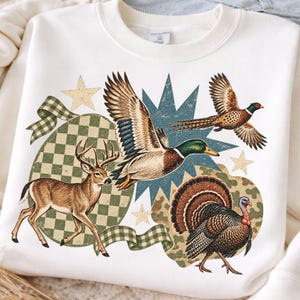 Peut inclure: Un sweat-shirt à col rond couleur crème arborant une impression graphique représentant un cerf, un canard colvert en vol, un faisan en vol et une dinde. Le motif inclut un damier vert, une étoile bleue et des étoiles beiges sur le tissu.