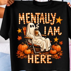 Peut inclure: Un t-shirt noir avec un motif représentant un fantôme blanc assis sur une chaise pliante, tenant une tasse de café. Le texte MENTALLY I AM HERE est écrit en lettres à carreaux orange et blanc. Le design inclut des citrouilles et des feuilles.