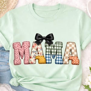 Peut inclure: T-shirt vert clair avec le mot "MAMA" en lettres patchwork colorées. Le "M" est en tricot rose, le "A" a un motif floral, le second "M" est à carreaux verts et bleus, et le dernier "A" est beige et orange. Un nœud noir est au-dessus.