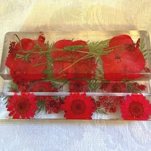 May include: Two rectangular, clear resin pieces with embedded red flowers and green foliage. The top piece has larger, round flowers, while the bottom piece features smaller, daisy-like blooms. The pieces are stacked, creating a layered effect.