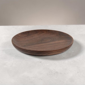 May include: A circular, dark brown wooden plate with a gently raised edge. The plate displays a natural wood grain pattern and is set against a neutral backdrop. This decorative plate is suitable for serving food or as a decorative item.