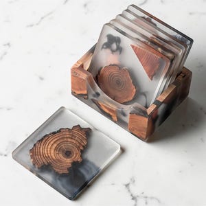 Puede incluir: Un juego de posavasos cuadrados con un diseño de madera encapsulado en resina transparente. Los posavasos se muestran en un soporte de madera. La madera tiene una veta natural y es de color marrón cálido. La resina tiene un aspecto ahumado y translúcido.