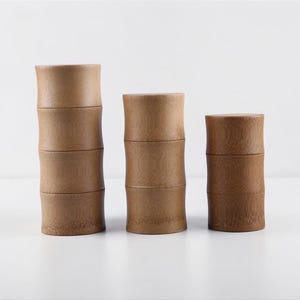 May include: Three stacks of bamboo containers. Each container is cylindrical with a slightly curved shape, resembling bamboo stalks. The containers are a light brown color and are stacked on top of each other. The background is white.