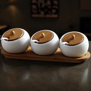 以下が含まれることがあります： 木製の蓋と小さなスプーンが付いた、白いセラミック製のスパイスジャー3つ。長方形の木製トレイにセットされています。ジャーは、テクスチャード加工されたドット模様です。蓋は丸く、小さなノブが付いています。