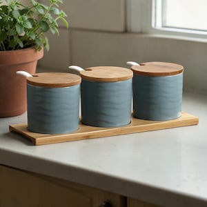 May include: Three matching blue ceramic canisters with wooden lids and small white spoons, set on a bamboo tray. The canisters have a wavy pattern and are arranged on a light-colored countertop, suggesting kitchen storage.
