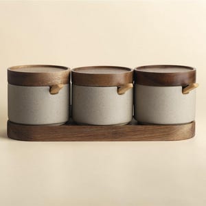以下が含まれることがあります： 木製の蓋と取っ手が付いた3つのセラミックキャニスターのセットで、木製のトレイに置かれています。キャニスターは薄いグレーで斑点のあるテクスチャで、蓋とトレイは温かみのある茶色です。