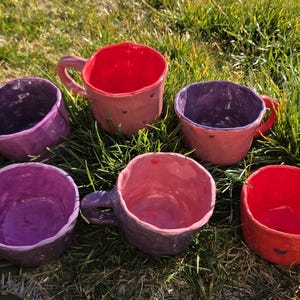 May include: Six handmade ceramic mugs in shades of red, pink, and purple are arranged on green grass. Each mug has a unique shape and texture, with a handle and a contrasting interior color. The mugs appear to be hand-painted.