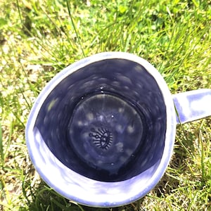 Puede incluir: Una taza de cerámica hecha a mano con interior y exterior azules. La taza tiene un asa y un diseño decorativo de ojo en la parte inferior. La taza está sobre hierba verde.