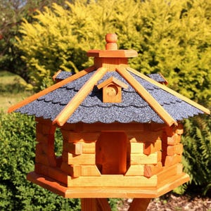 May include: A wooden birdhouse with a gray shingled roof and a small decorative chimney. The birdhouse is constructed from light-colored wood and features a front opening and a small bird-shaped cutout. It is mounted on a wooden stand.