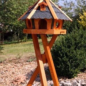 May include: A wooden birdhouse with a peaked roof and a stand. The birdhouse has a natural wood finish with a dark gray shingled roof. The stand is made of wood and has a cross-legged design. The birdhouse has round entry holes.