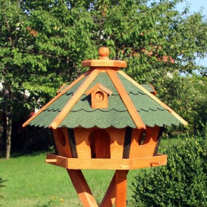 May include: A wooden birdhouse with a green shingled roof and a decorative finial. The birdhouse has multiple openings and is mounted on a wooden stand. The birdhouse is made of natural wood and is designed to attract birds to a garden.