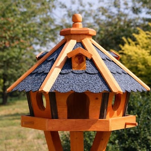 Könnte beinhalten: Ein hölzernes Vogelhaus mit mehrstöckigem Design. Das Dach ist mit dunkelblauen Schindeln bedeckt und wird von hellbraunen Holzbalken getragen. Das Vogelhaus hat eine runde Öffnung und eine kleine Sitzstange. Die Struktur besteht aus hellbraunem Holz.