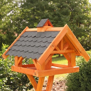 May include: A wooden bird feeder with a peaked roof and a small chimney. The roof is covered in gray shingles, and the feeder is constructed from light-colored wood. The feeder is supported by a wooden stand and is set outdoors in a garden.
