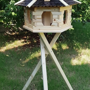 Op de afbeelding: Een houten vogelhuisje met een donkerbruin pannendak en een lichtgekleurd houten frame. Het vogelhuisje heeft meerdere openingen en staat op een lichtgekleurde houten standaard met een X-vormige basis. Het vogelhuisje staat in een grasrijke buitenomgeving.