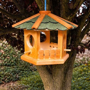 May include: A wooden birdhouse with a green shingled roof and a hexagonal shape. The birdhouse has a round entry hole and a small wooden perch. It is suspended from a blue rope and mounted on a tree.